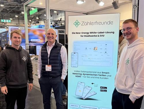 Drei Männer stehen auf einem Messestand vor einem türkisfarbenen Roll-up-Banner der Zählerfreunde GmbH. In der Mitte lächelt Professor Jens Bockstette, gekleidet in Hemd und dunkler Steppweste. Links neben ihm steht Jakob Kungel in einem dunklen Hoodie mit Firmenlogo, rechts Tobias Keussen in einem hellen Hoodie mit demselben Logo. Das Banner bewirbt eine „New-Energy White-Label-Lösung für Stadtwerke & EVU“ und zeigt eine App-Oberfläche zur Visualisierung von Smart-Metering- und Energiedaten. Im Hintergrund sind weitere Messestände und Hallenbeleuchtung zu sehen.